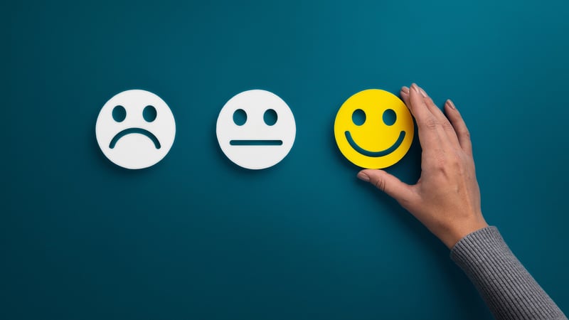 Ein trauriger, neutraler und fröhlicher Smiley nebeneinander, der fröhliche Smiley ist gelb und wird von einer Hand gehalten